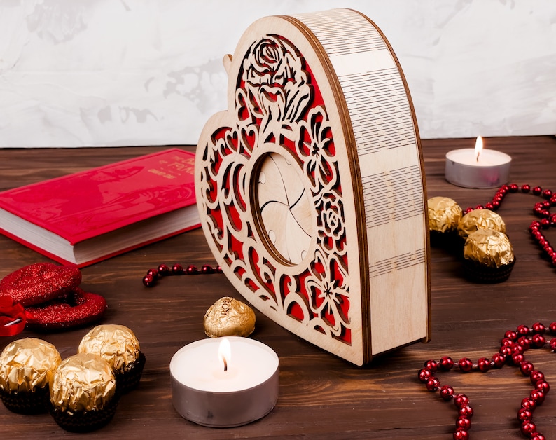 May include: A wooden heart-shaped box with a red floral design and a cutout in the center. The box is surrounded by red beads, gold-wrapped chocolates, and two white candles.