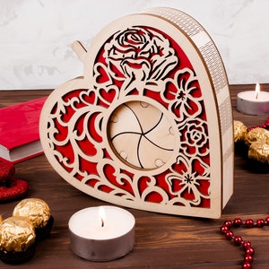 May include: A wooden heart-shaped box with a red and white floral design. The box has a cutout in the center and is decorated with a rose and other flowers. The box is sitting on a wooden surface with candles and chocolates.
