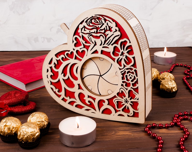 May include: A wooden heart-shaped box with a red and white floral design. The box has a cutout in the center and is decorated with a rose and other flowers. The box is sitting on a wooden surface with candles and chocolates.