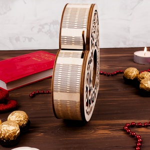 May include: A wooden heart-shaped box with a decorative design. The box is open and has a red book and chocolate candies next to it. There are also red beads and a lit candle on a wooden surface.