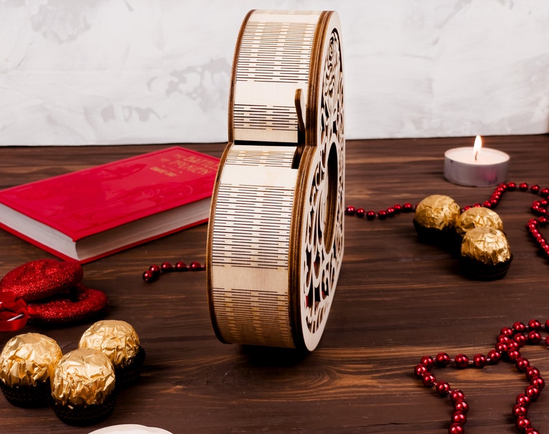 May include: A wooden heart-shaped box with a decorative design. The box is open and has a red book and chocolate candies next to it. There are also red beads and a lit candle on a wooden surface.