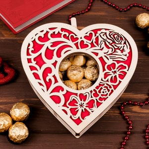 May include: A wooden heart-shaped box with a red and white floral design. The box has a cutout in the center that reveals a collection of gold-wrapped chocolates.