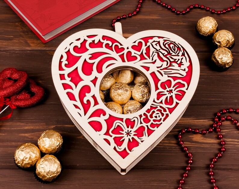 May include: A wooden heart-shaped box with a red and white floral design. The box has a cutout in the center that reveals a collection of gold-wrapped chocolates.