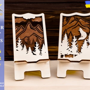 May include: Two wooden phone stands with a mountain and forest design. The stand on the left has a mountain scene, while the stand on the right has a mountain scene with a wolf silhouette.