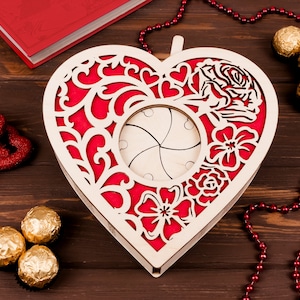 May include: A wooden heart-shaped box with a red and white floral design. The box has a hinged lid and a cutout in the center. The box is surrounded by red beads and chocolate candies.