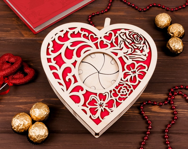 May include: A wooden heart-shaped box with a red and white floral design. The box has a hinged lid and a cutout in the center. The box is surrounded by red beads and chocolate candies.