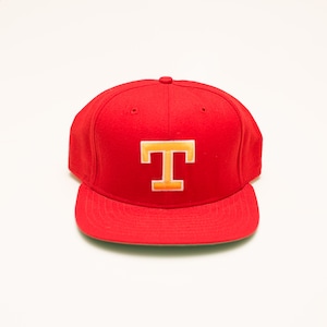 Puede incluir: Gorra de béisbol roja con visera plana. La gorra presenta una letra "T" bordada en oro y naranja en la parte delantera. Este sombrero es de estilo clásico, adecuado para uso informal.