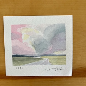 May include: Watercolor landscape painting featuring a winding road through a green field under a dramatic sky. The sky blends pink, yellow, and gray hues. The artwork is signed and dated "2025".