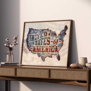 Puede incluir: Una impresión de estilo vintage del mapa de los Estados Unidos de América con las palabras "The United States of America Est. 1776" en una fuente colorida y desgastada. La impresión está enmarcada en un marco de madera marrón claro y está colgada en una pared blanca.