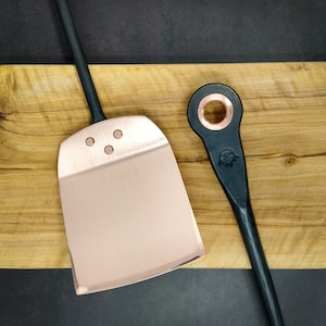 May include: A copper pizza peel with a black wooden handle and a black metal pizza cutter with a copper ring on a wooden chopping board.