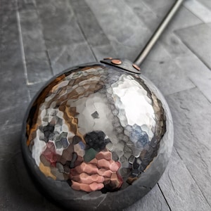 Peut inclure: Une louche en m&eacute;tal martel&eacute; avec un long manche. Le bol rond est textur&eacute; et r&eacute;fl&eacute;chissant. La poign&eacute;e sombre est orn&eacute;e de d&eacute;tails en cuivre, sugg&eacute;rant un ustensile de cuisine rustique et artisanal, id&eacute;al pour cuisiner.