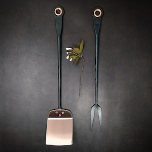May include: A set of two black metal kitchen utensils with copper accents. The utensils include a spatula and a fork.