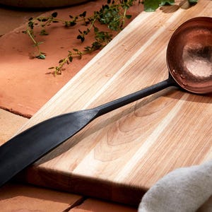 May include: A black handled copper ladle with a hammered finish sits on a wooden cutting board.