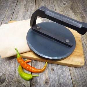 Peut inclure: Une presse à tortillas en fonte noire sur une planche à découper en bois avec deux piments.