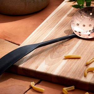 May include: A black handled copper slotted spoon with a wooden cutting board and pasta.