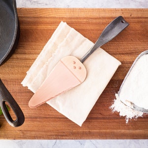 May include: A wooden cake server with a copper-coloured blade and a black handle. The server is lying on a white cloth on a wooden chopping board. There is a cast iron skillet and a scoop of flour in the background.