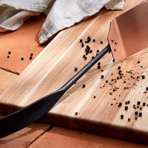 May include: A black handled copper pizza peel on a wooden chopping board with black peppercorns scattered on the surface.