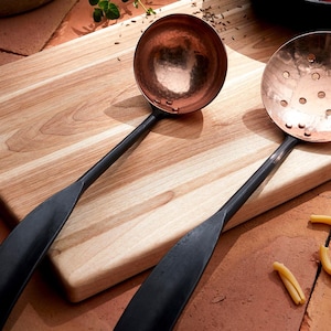 May include: Two copper ladles with black handles on a wooden chopping board. One ladle has a smooth bowl and the other has a perforated bowl.