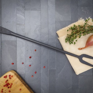 May include: A black metal grilling fork with a copper-colored handle. The fork is lying on a slate surface with red peppercorns scattered around it. A small bunch of thyme and a red onion are on a beige cloth napkin.