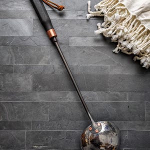 Peut inclure: Une louche en m&eacute;tal fabriqu&eacute;e &agrave; la main, avec un manche en bois fonc&eacute; et des accents de cuivre. La louche pr&eacute;sente une cuill&egrave;re sph&eacute;rique martel&eacute;e et un long manche fin. Une boucle en cuir est attach&eacute;e &agrave; la poign&eacute;e.