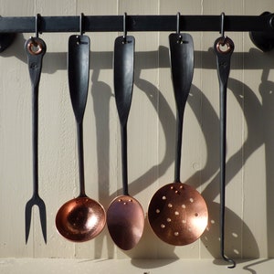 Peut inclure: Un ensemble de six ustensiles de cuisine en métal noir avec des têtes en cuivre suspendus à un support en métal noir. Les ustensiles comprennent une fourchette, une louche, une cuillère, une cuillère à fentes et deux autres outils.