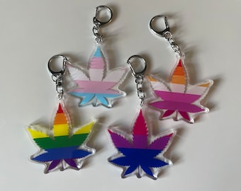 Puede incluir: Cuatro llaveros de acrílico con forma de hojas de marihuana, cada uno con un esquema de color diferente que representa una bandera del orgullo. Las banderas son: transgénero, lesbiana, bisexual y gay.