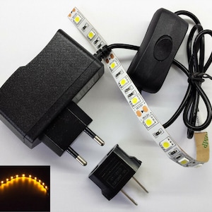 May include: A black power adapter, a black on/off switch, and a strip of LED lights with a DC12V marking. The LED strip is flexible and has a warm white glow. A smaller image shows the LED strip lit up.