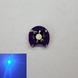 May include: A small, round, purple circuit board with a white LED light in the center. The circuit board has four silver contacts on the side.
