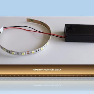 Może przedstawiać: Pasek LED o ciepłym białym świetle z czerwonymi i czarnymi przewodami podłączonymi do czarnego akumulatora. Pasek LED jest zwinięty w kółko, a akumulator znajduje się na białej powierzchni.