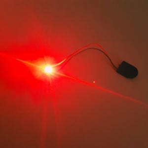 Puede incluir: Una luz LED roja brillante con un haz enfocado, que emana de un punto central. La luz está conectada a una pequeña batería negra a través de finos cables rojos y negros. El fondo es un degradado de rojo.