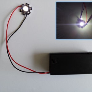 Puede incluir: Una pequeña luz LED blanca con dos cables conectados a una batería negra. La luz está encendida y brilla intensamente.