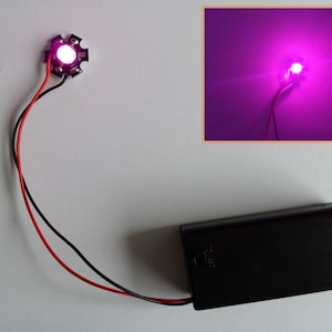 Puede incluir: Una luz LED rosa con dos cables conectados a una batería negra. El LED está encendido y proyecta una luz rosa brillante.