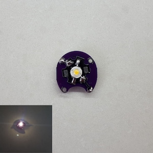 Könnte beinhalten: Eine kleine, runde, lila Leiterplatte mit einer hellweißen LED in der Mitte. Die Platine hat mehrere kleine schwarze Komponenten und silberne Lötstellen. Ein kleineres Bild zeigt die beleuchtete LED.