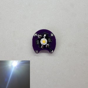 May include: A small, round, purple circuit board with a white LED light in the center. The LED is surrounded by four silver wires.