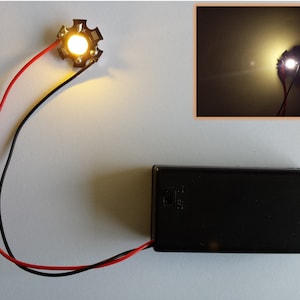 Puede incluir: Una pequeña luz LED redonda blanca con dos cables conectados a ella. La luz está conectada a una batería negra. La luz está encendida e ilumina un fondo oscuro.