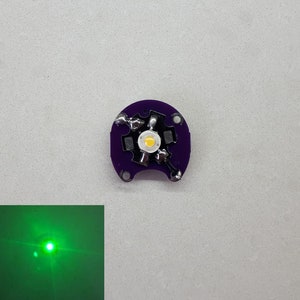 以下が含まれることがあります： 中央に白いLEDライトが付いた、小さく丸い紫色の回路基板。LEDは明るい緑色の光を放っています。