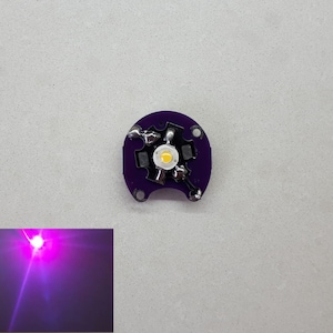 Peut inclure: Un petit circuit imprimé violet avec une LED blanche au centre. La LED émet une lumière violette brillante.