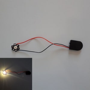 May include: A small, round, white LED light with a black circuit board and two wires connected to a black battery pack.