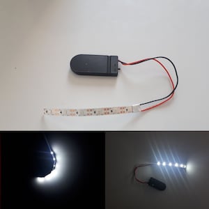 May include: A black battery pack with red and black wires connected to a strip of white LED lights. The lights are turned on and are illuminating a dark surface.