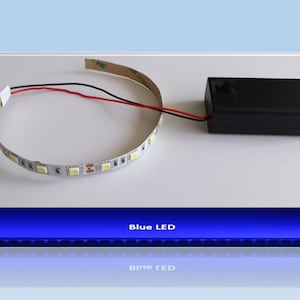 Puede incluir: Una tira de luz LED blanca con un cable negro y un cable rojo conectados a una batería negra. El texto "Blue LED" es visible debajo de la imagen.