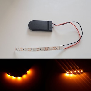 Könnte beinhalten: Ein LED-Lichtstreifen mit einem Akkupack und Kabeln. Die Lichter sind orange und in der zweiten Abbildung eingeschaltet.