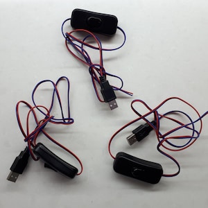 Puede incluir: Tres interruptores USB en línea negros con cables rojos y azules. Cada interruptor tiene una carcasa de plástico negro y un pequeño botón negro.