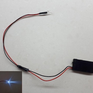 Może przedstawiać: Mała biała dioda LED z czarnymi przewodami i pakietem baterii. Światło jest włączone i oświetla białą powierzchnię.