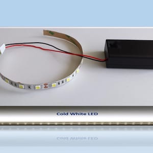 Puede incluir: Una tira de luces LED blancas frías con una batería negra. La tira LED está enrollada y tiene cables rojos y negros.
