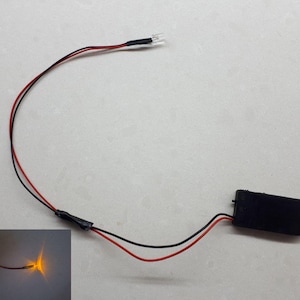 Op de afbeelding: Een klein, batterijgevoed LED-lampje met een zwarte batterijhouder en rode en zwarte draden. Het lampje is aan en geeft een heldere gele gloed af.