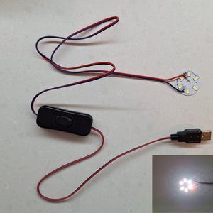 Puede incluir: Una luz LED blanca con un conector USB y un interruptor en línea. La luz está conectada a un cable negro con cables rojo y azul. La luz está encendida e iluminando una superficie blanca.