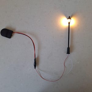 Könnte beinhalten: Eine Miniatur-Straßenlaterne in Schwarz mit warmem, leuchtendem Licht. Die Lampe ist über rote und weiße Drähte mit einem schwarzen Akku verbunden. Der Laternenpfahl ist schwarz mit einem dekorativen Aufsatz.