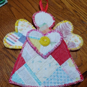 May include: A handmade patchwork angel ornament with a red heart and a yellow button. The angel's wings are made of different fabrics with yellow trim.