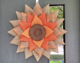 May include: A brown, orange, and tan burlap sunflower wreath hanging on a black door. The wreath is made of burlap and has a brown center.