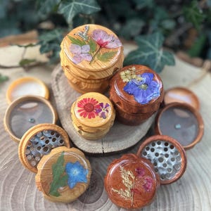 May include: A collection of wooden herb grinders with floral designs. The grinders feature pressed flowers and resin on the lids, with various colors and shapes. The grinders are displayed on a wooden surface.